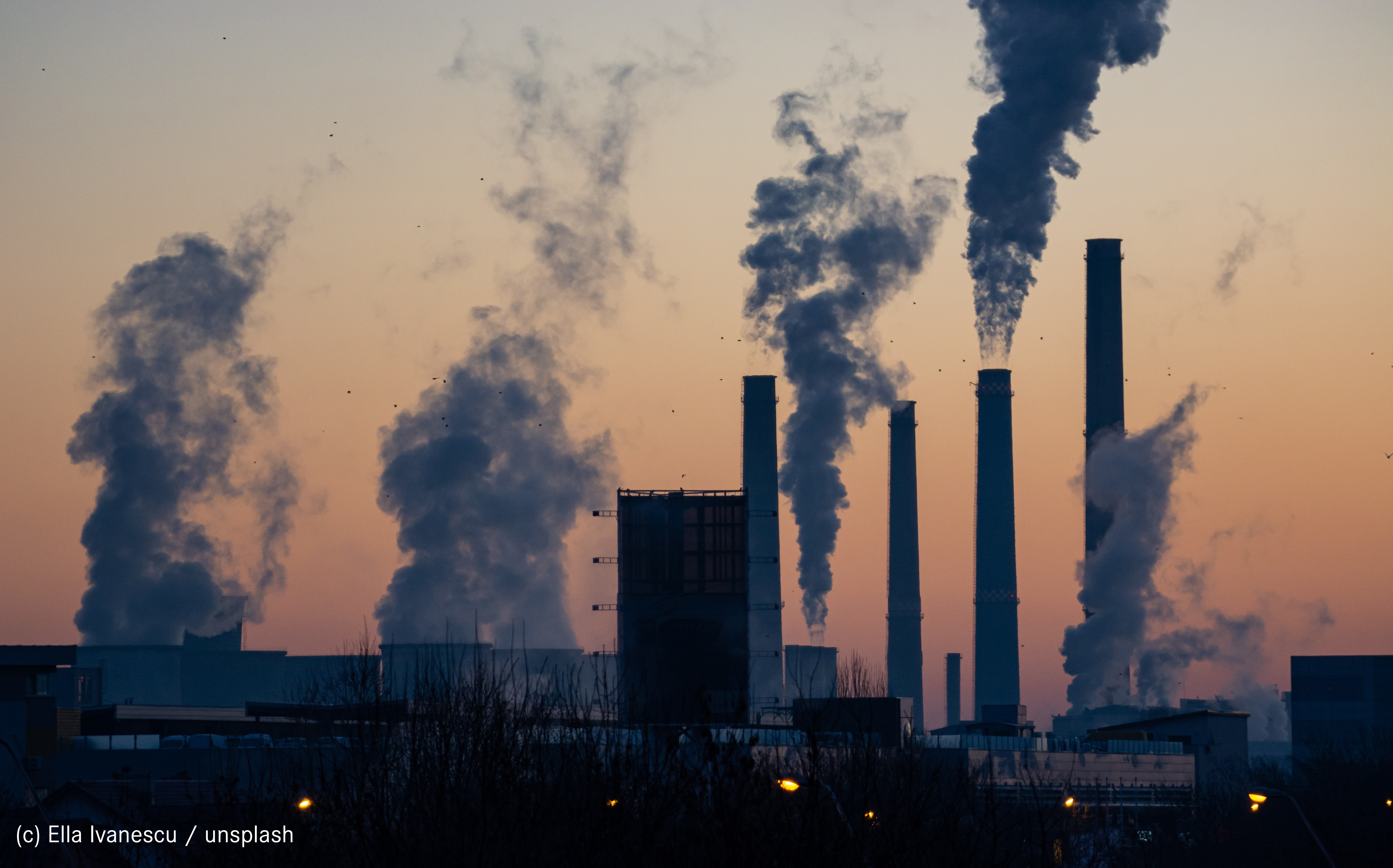 Fabrik im Sonenuntergang mit mehreren Türmen aus denen Rauch kommt.