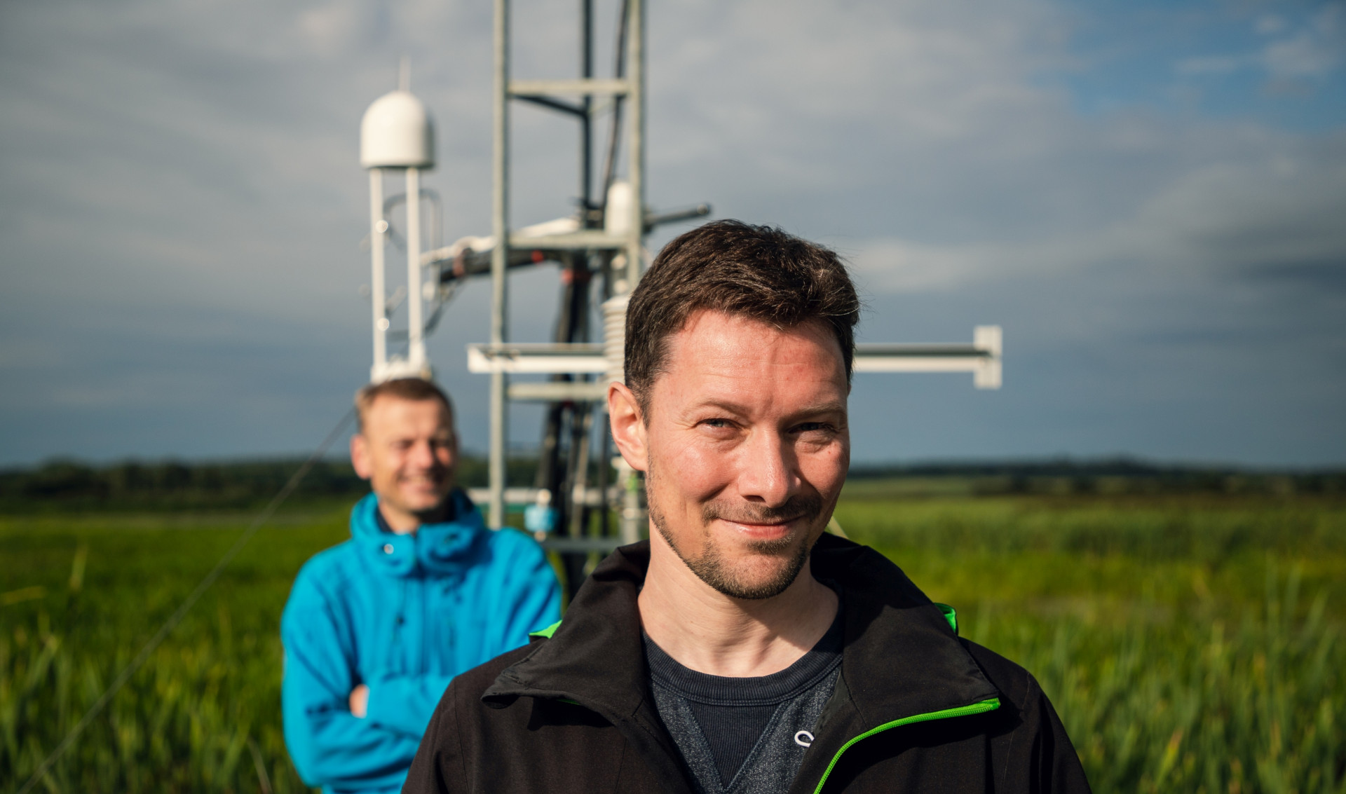 Two men are standing in front of a small tower with sensors in a field full of green plants.