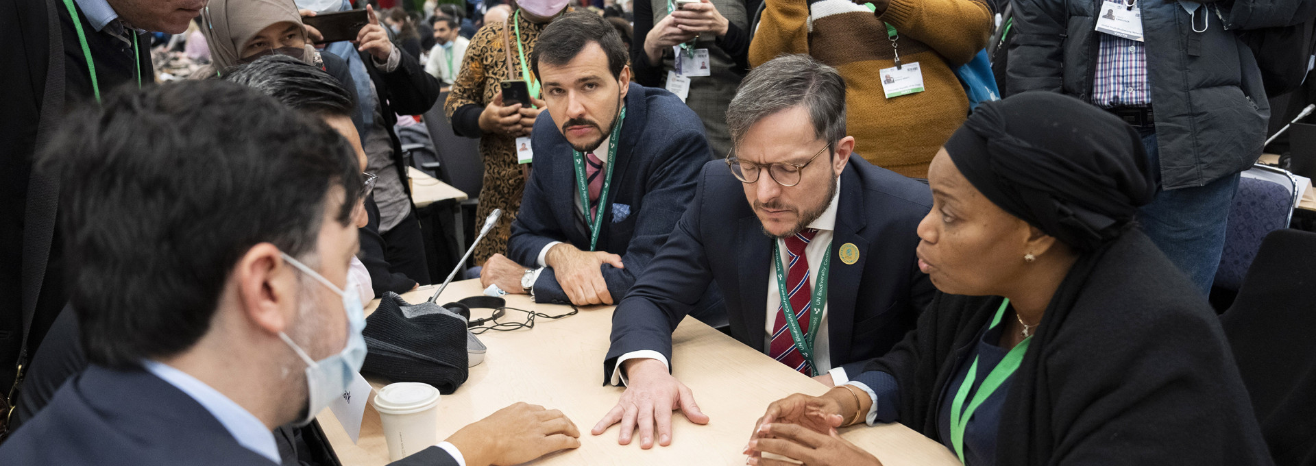 COP15 participants from different countries sitting at a table negotiating, and being photographed from people standing in the background.