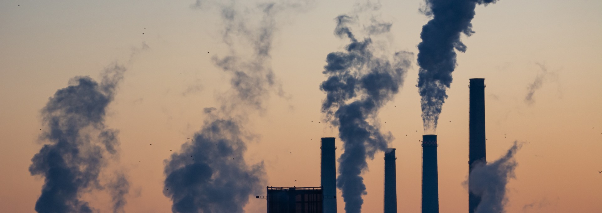 Fabrik im Sonenuntergang mit mehreren Türmen aus denen Rauch kommt.
