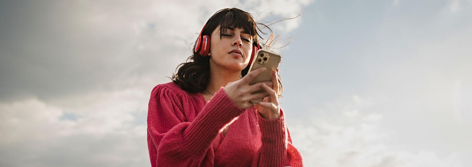 Eine Person mit braunen Haaren und Kopfhörern am Smartphone vor wolkigem Himmel