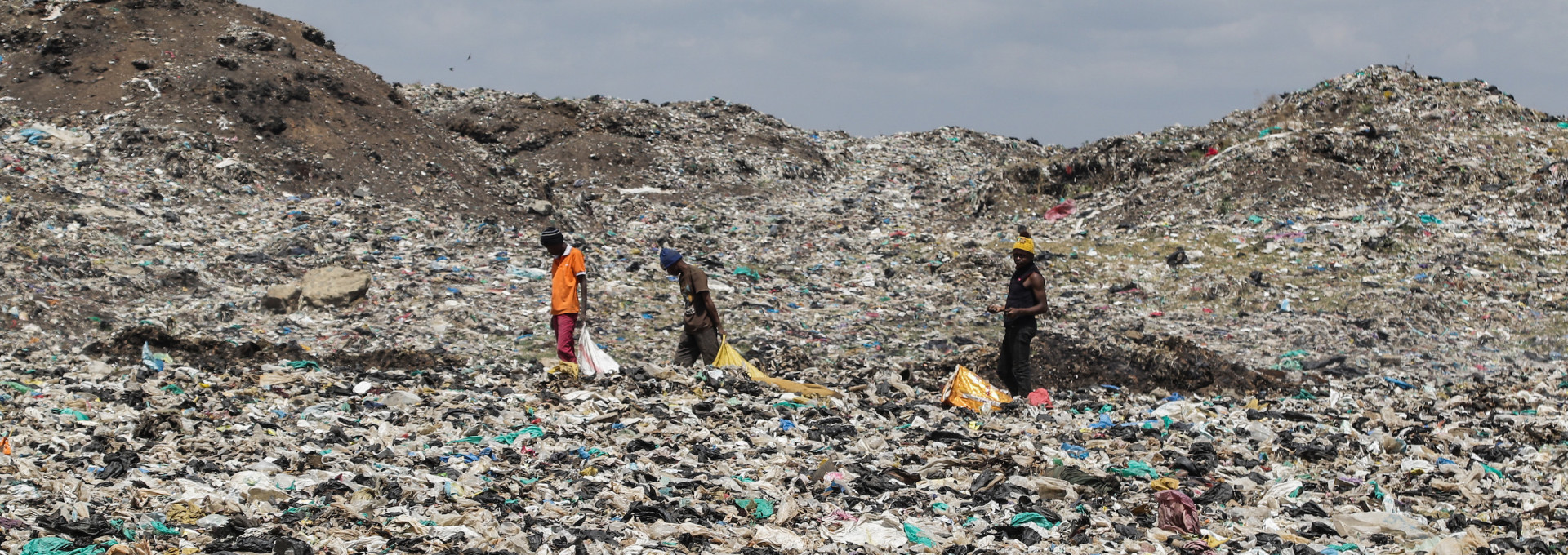 Drei Personen laufen über eine riesige Müllhalde mit Plastik und anderem Müll