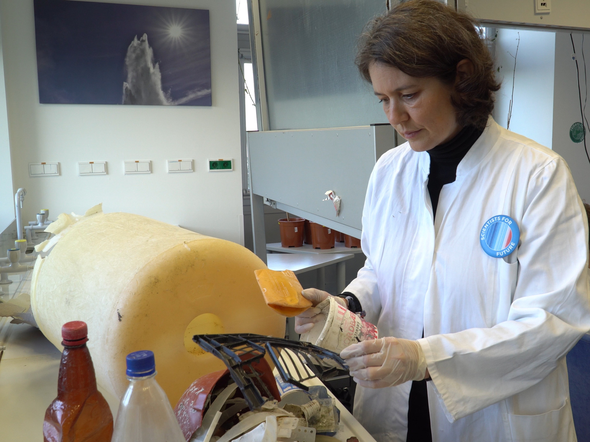 Wissenschaftlerin im weißen Kittel mit einem großen gelben Stück Plastikmüll im Labor