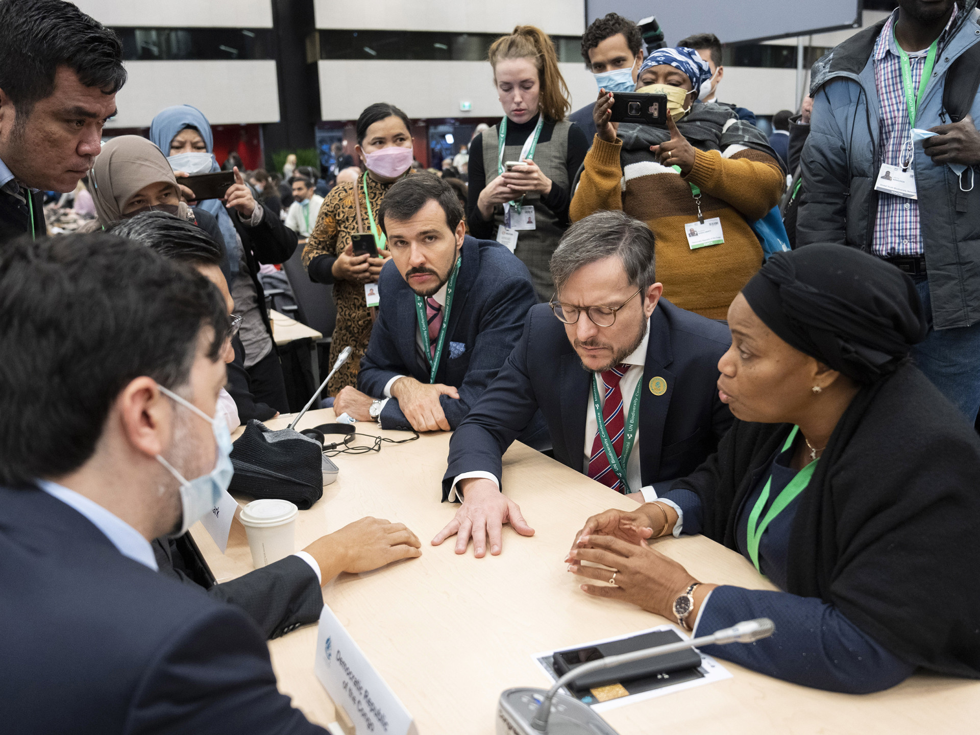 COP15 participants from different countries sitting at a table negotiating, and being photographed from people standing in the background.