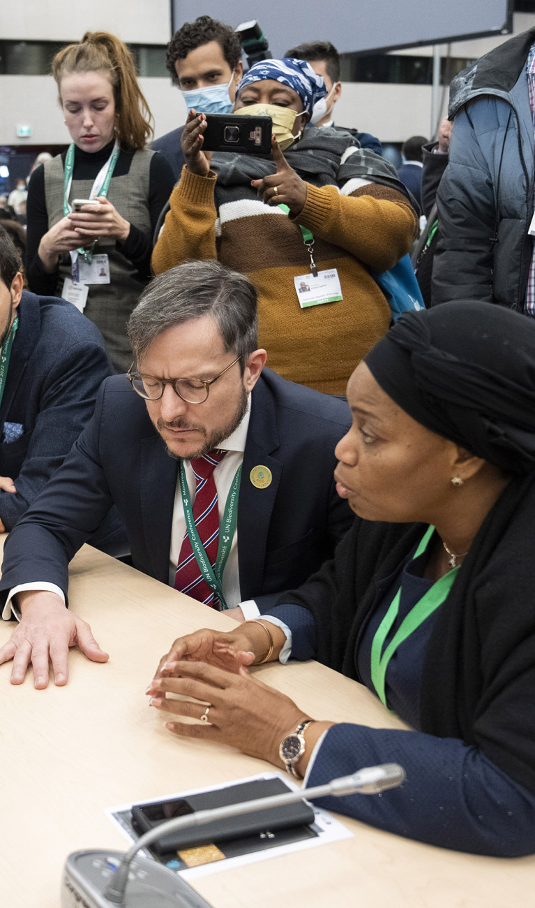 Teilnehmende der UN-Biodiversitätskonferenz verschiedener Länder sitzen an einem Tisch und unterhalten sich, im Hintergrund machen andere Fotos. Sie schauen angespannt.