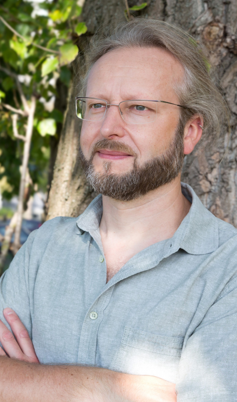 Portrait Björn Rau steht vor Baum, trägt ein grünes Hemd. Arme verschränkt.