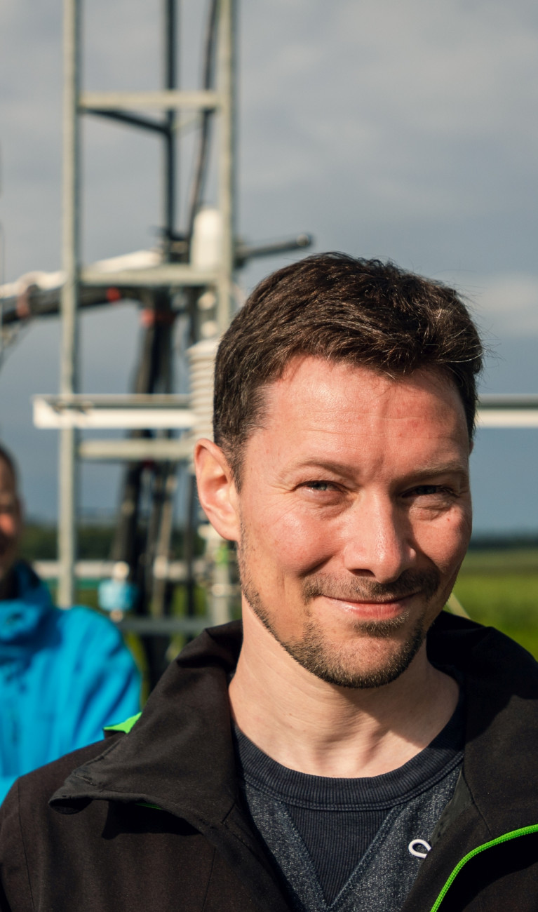 Two men are standing in front of a small tower with sensors in a field full of green plants.