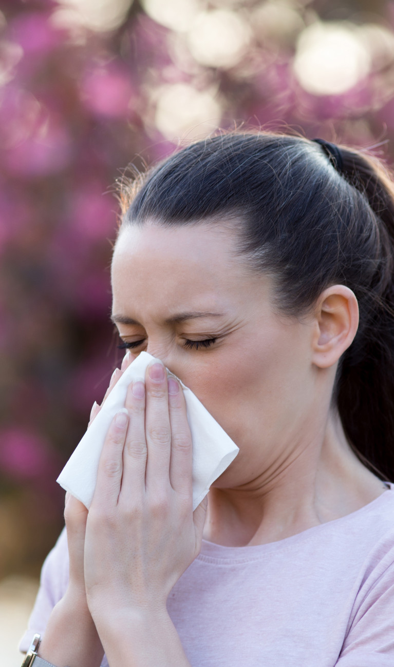 A person blowing their nose, with flowers and trees in the background