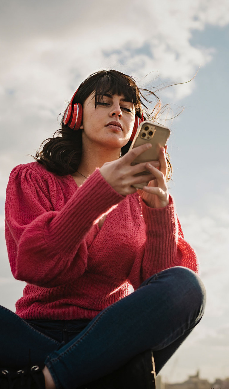 Eine Person mit braunen Haaren und Kopfhörern am Smartphone vor wolkigem Himmel