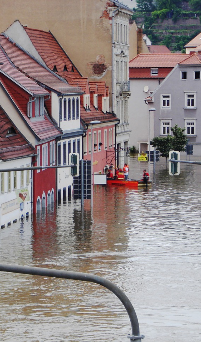 Eine Straße steht komplett unter Wasser, Menschen in einem Schlauchboot retten Personen aus einem Haus