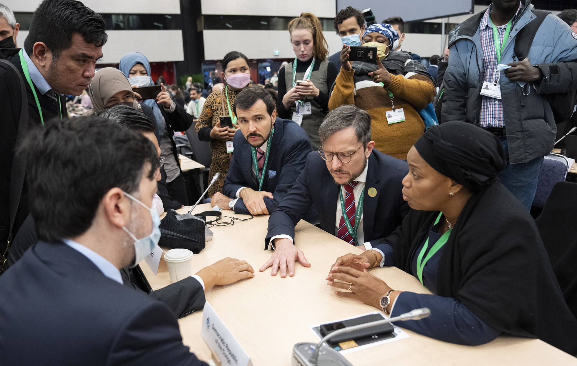 COP15 participants from different countries sitting at a table negotiating, and being photographed from people standing in the background.