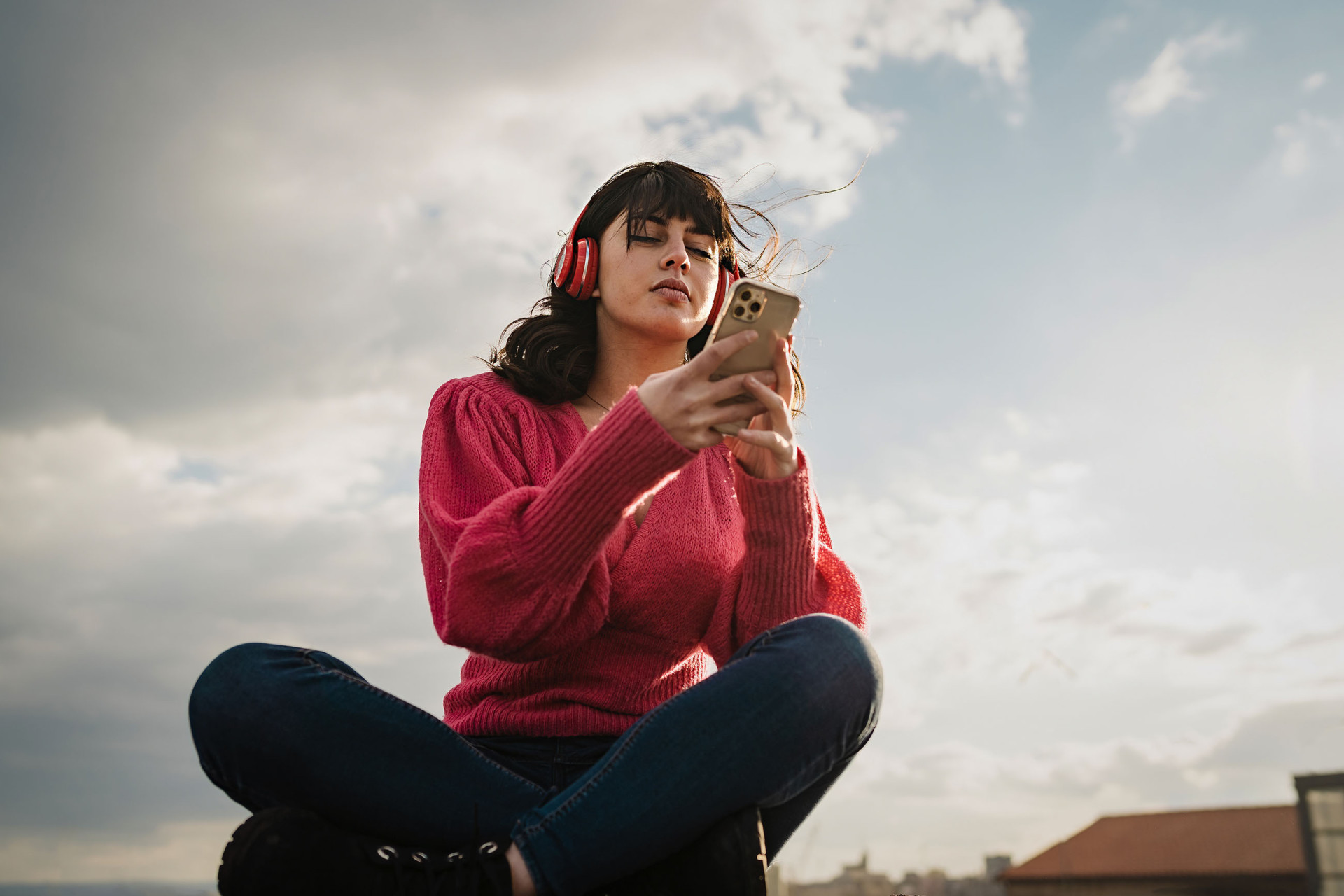 Eine Person mit braunen Haaren und Kopfhörern am Smartphone vor wolkigem Himmel