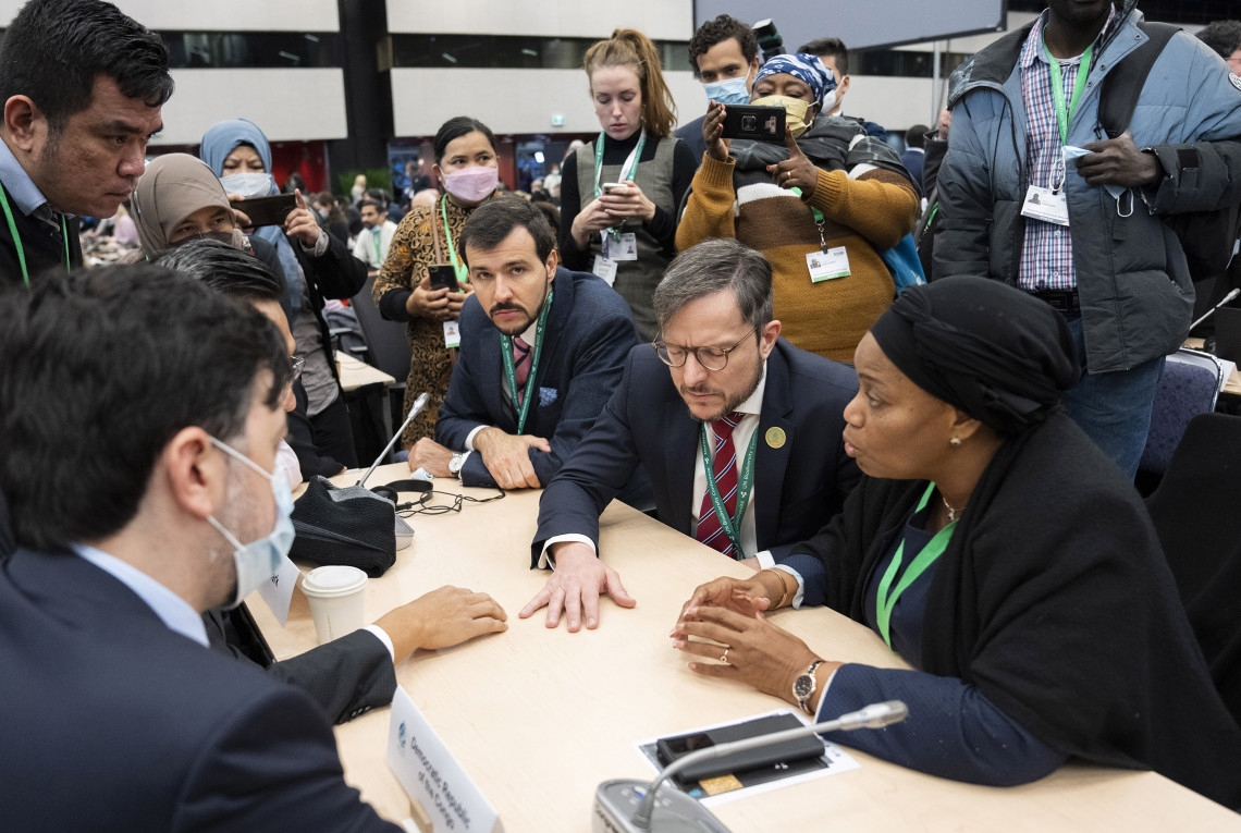 Teilnehmende der UN-Biodiversitätskonferenz verschiedener Länder sitzen an einem Tisch und unterhalten sich, im Hintergrund machen andere Fotos. Sie schauen angespannt.