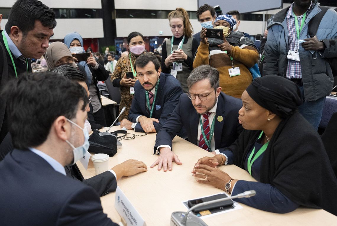 COP15 participants from different countries sitting at a table negotiating, and being photographed from people standing in the background.