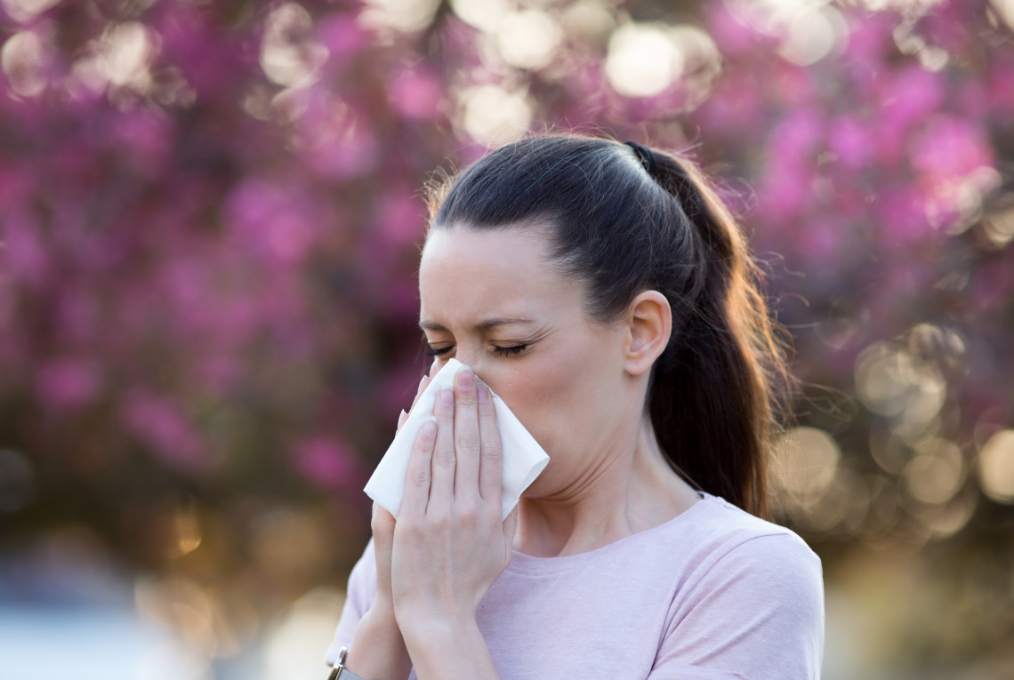Pollen Eine Person schneutzt sich die Nase, dahinter blüht ein Baum.