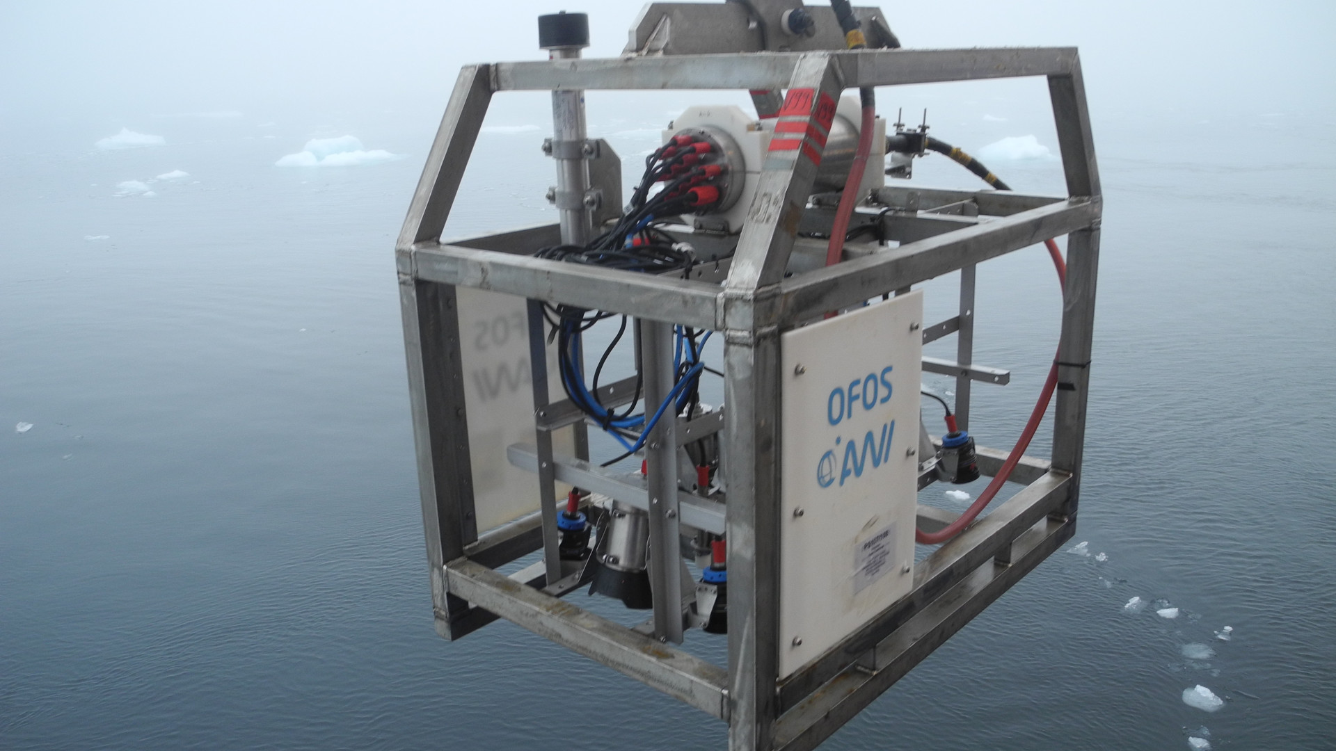 Wissenschaftliches Gerät in einem Metallkäfig schwebt über dem Wasser, auf dem kleine Eisschollen treiben